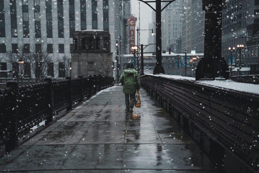 Image of a person with a green coat and a tan bag walking across a bridge crossing the Chicago river while the snow is falling