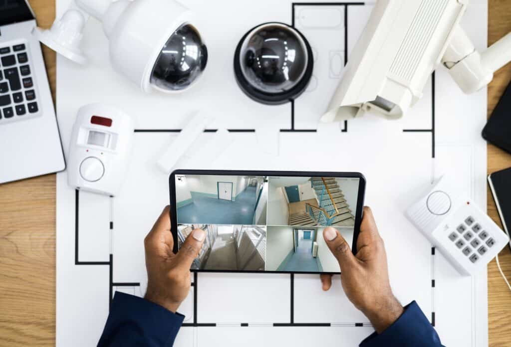 Image of blueprint with security cameras resting on top of them and a man holding a tablet looking at a security feed